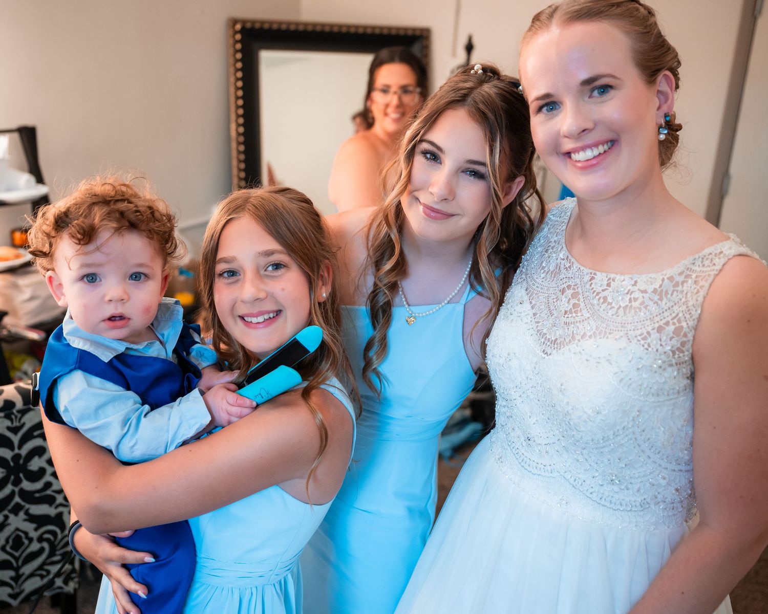 Four children in blue formal attire smile together indoors, with a woman visible in a mirror behind them.