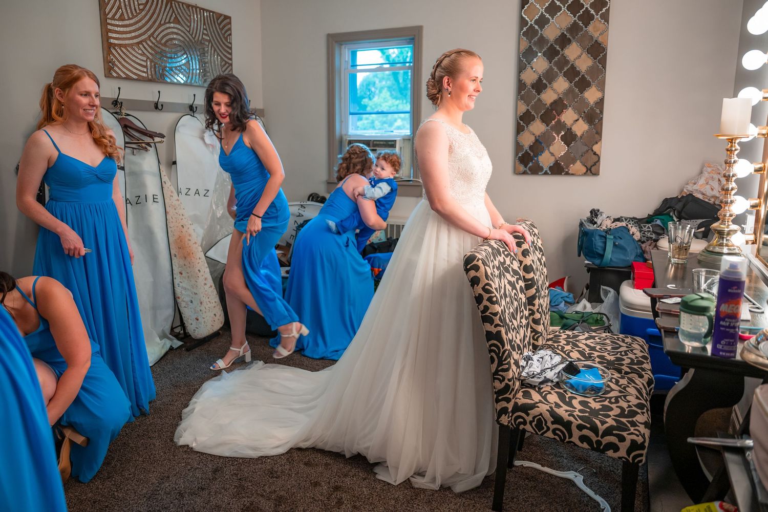 Bridesmaids in blue dresses help a bride in a white gown prepare in a bridal suite before the wedding.