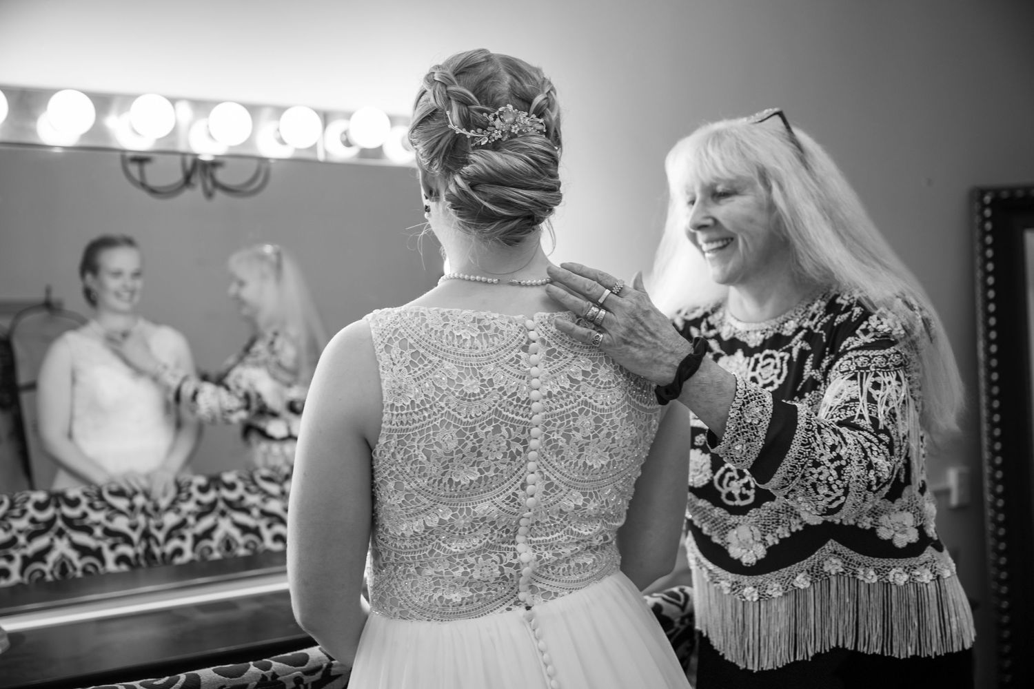 Mother helps bride with beaded wedding gown back detail in bridal suite mirror.