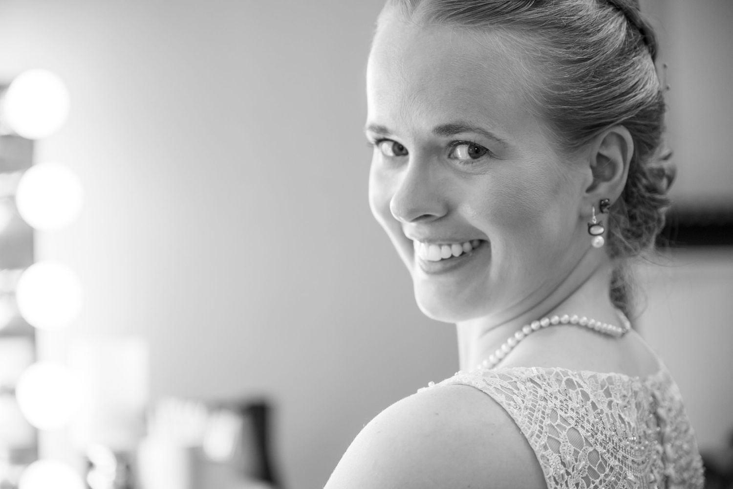 Black and white portrait of a smiling woman wearing pearl jewelry and a lace dress near a vanity mirror.
