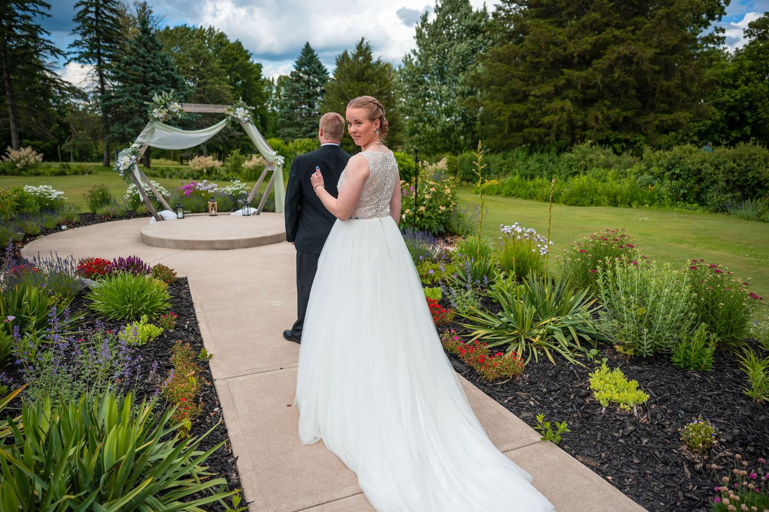 Bride in flowing white gown poses with groom in lush garden wedding venue with floral arch backdrop.