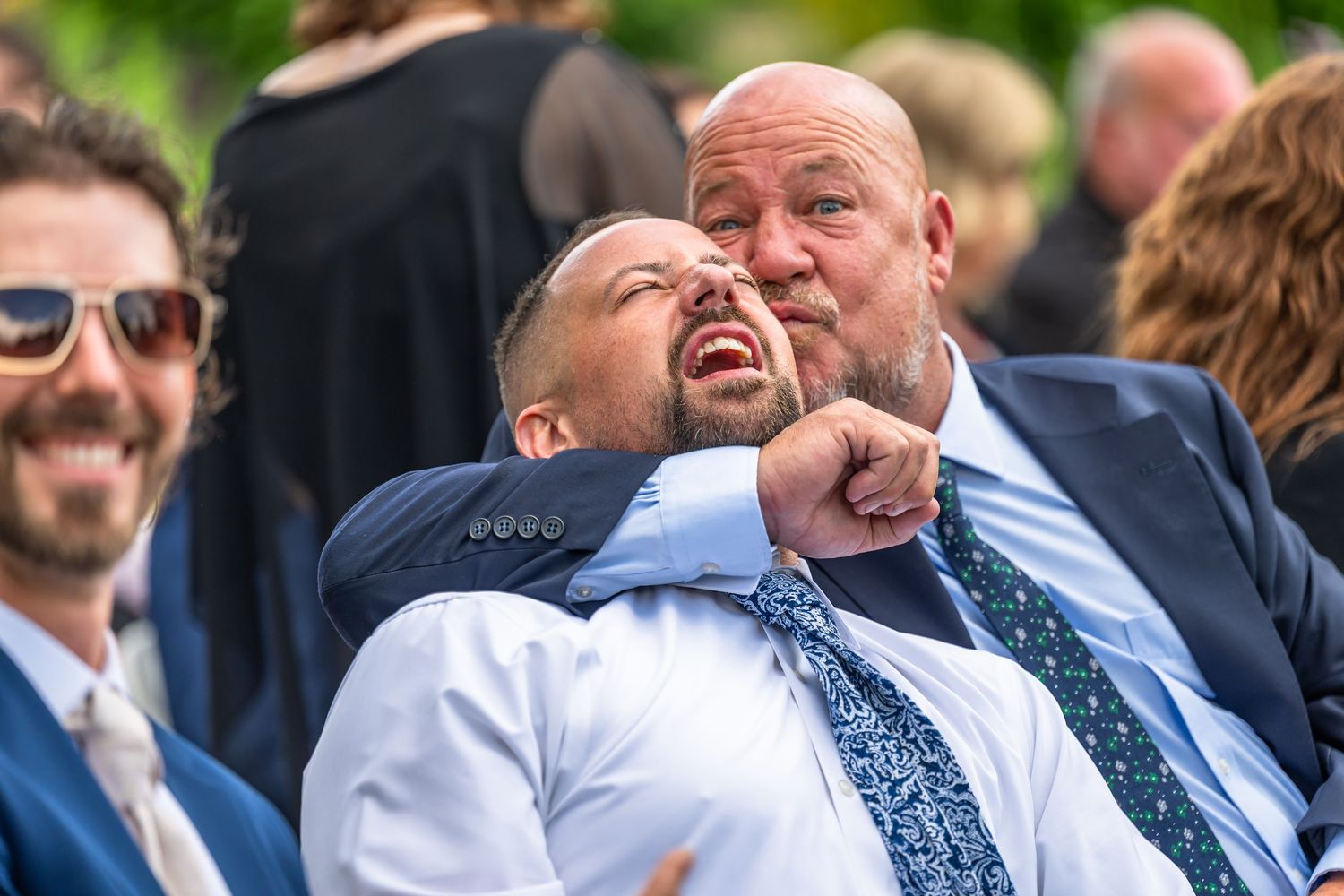 Two men laughing joyfully while seated as wedding guests during an outdoor wedding ceremony.