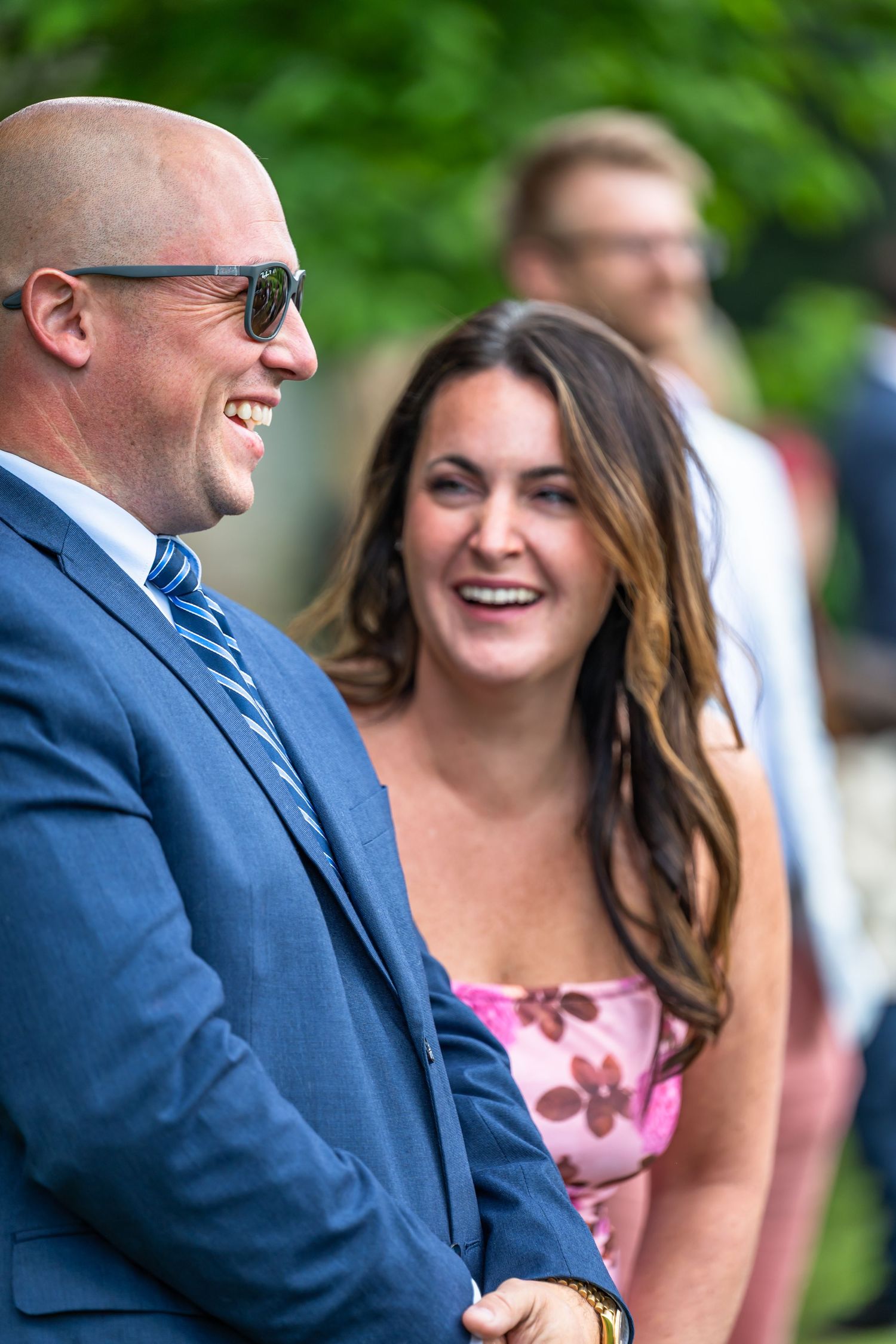 A bald man in a blue suit and sunglasses laughs with a smiling woman in a pink floral dress at a wedding.