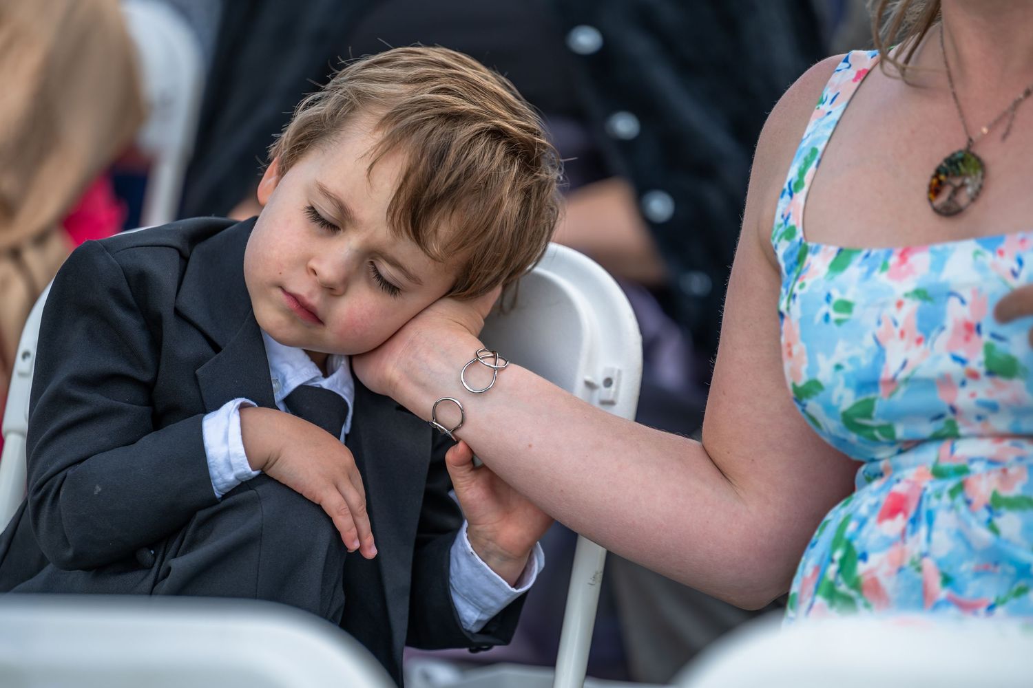 Tired young boy in suit rests head on tattooed arm of woman at outdoor wedding ceremony.