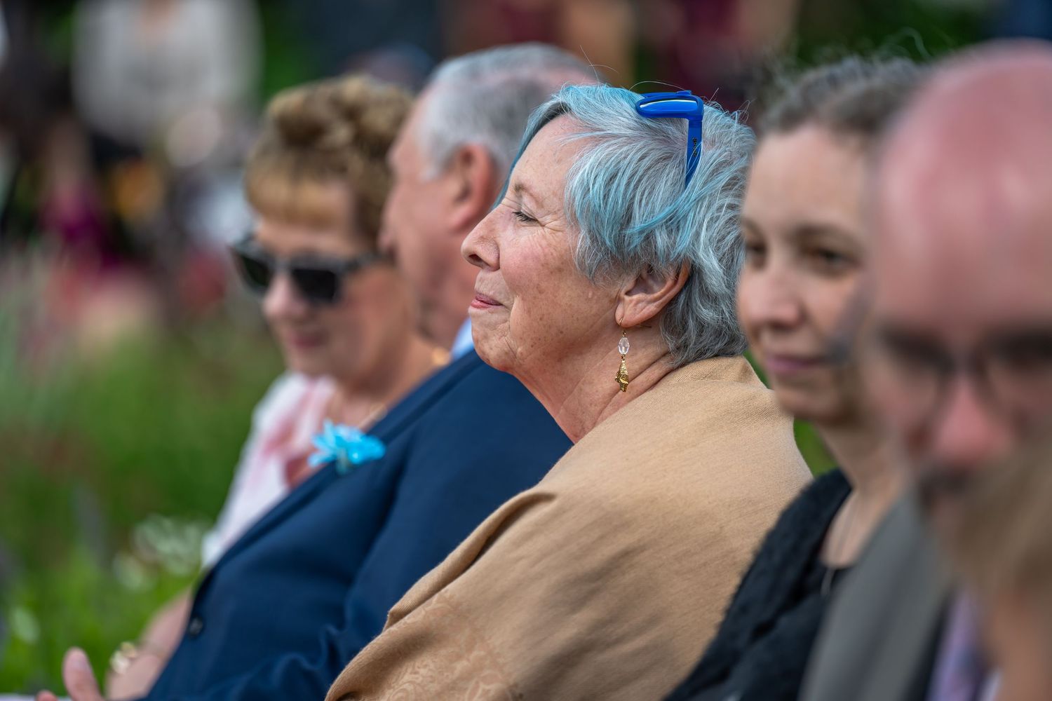 Smiling wedding guest with blue-gray hair enjoys outdoor ceremony seated among other guests.