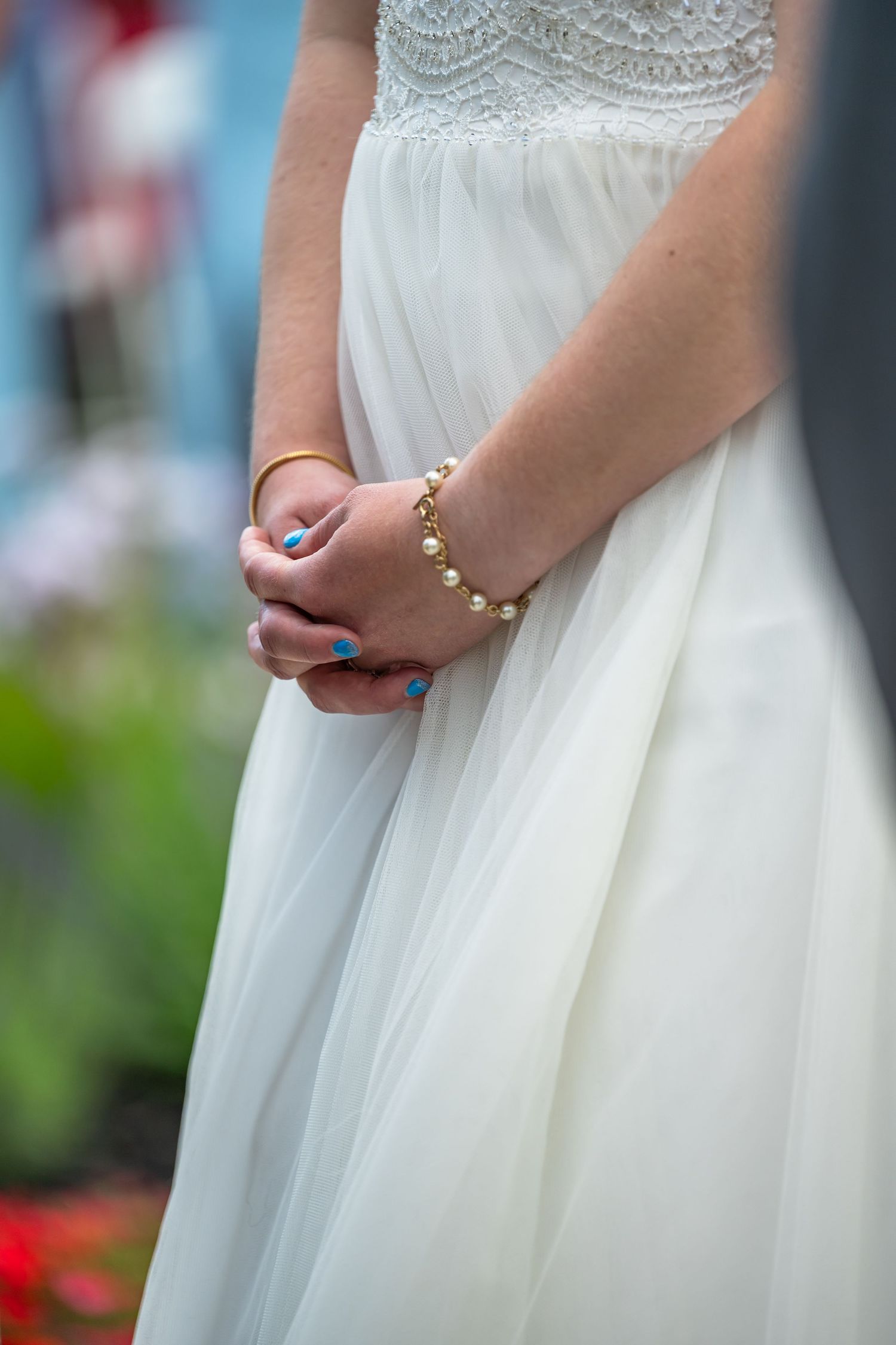 Bride in white wedding dress clasping hands showing pearl bracelet and blue nail polish.