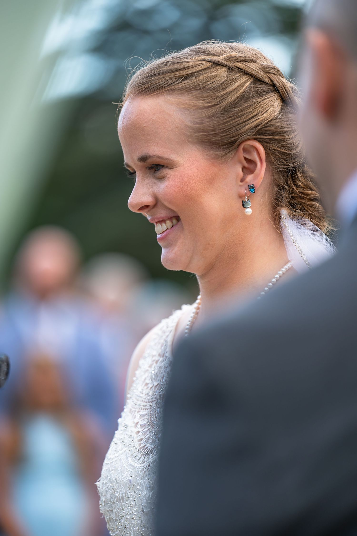 Smiling bride with braided updo and teal earrings during outdoor wedding ceremony.