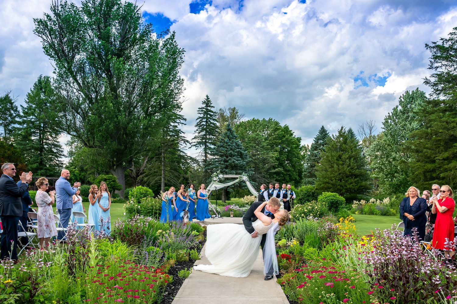 Bride and groom share a romantic dip kiss during outdoor garden wedding ceremony with bridesmaids in blue dresses.