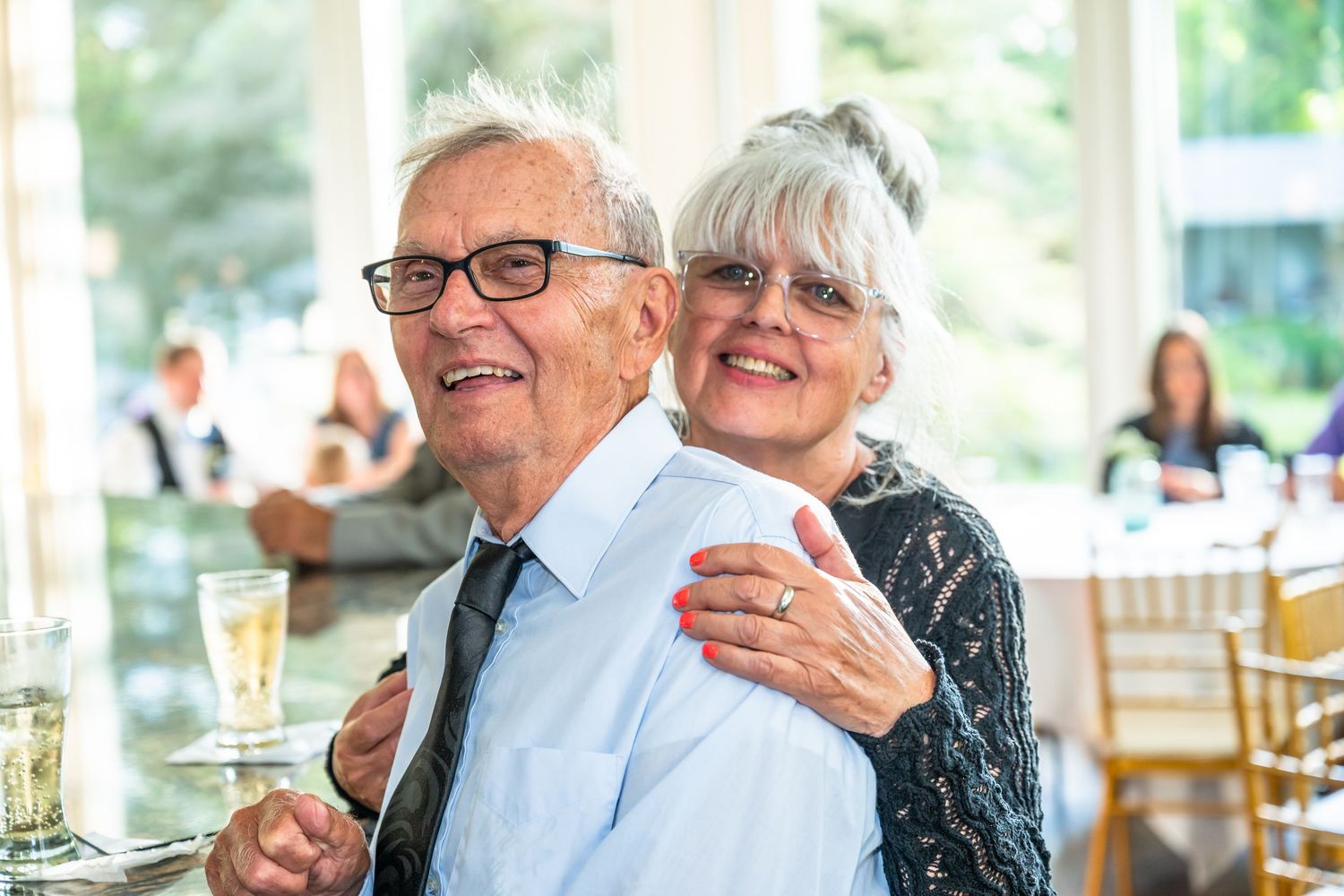 Smiling older couple embracing at a wedding reception in a bright sunlit venue.