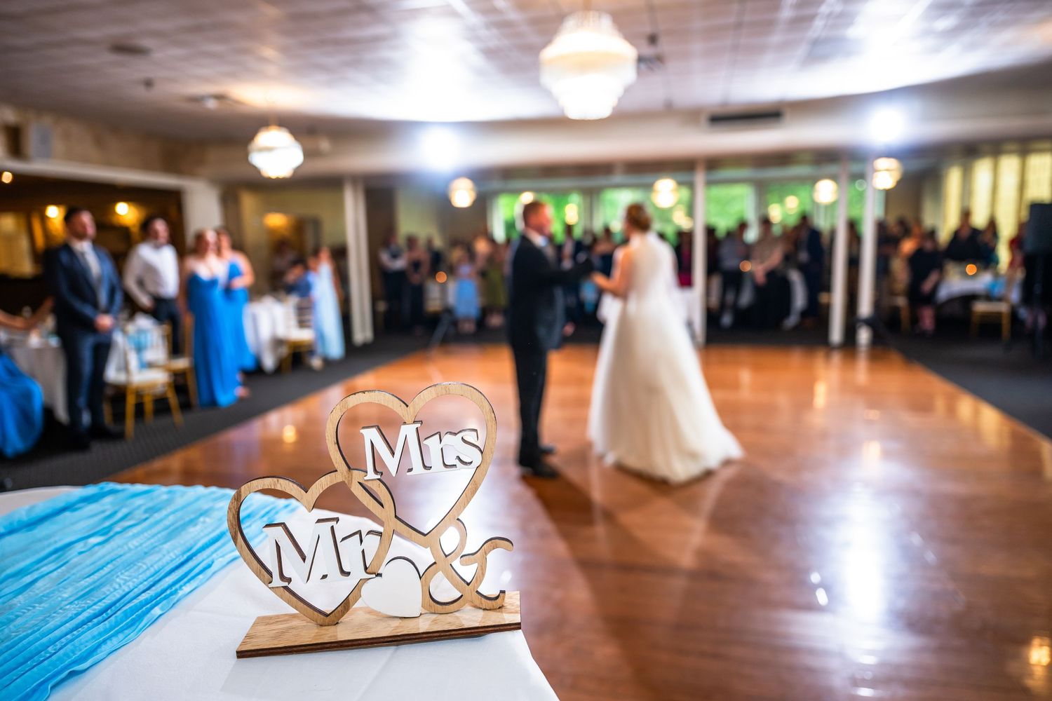 Mr and Mrs wooden sign on a table with newlyweds sharing their first dance at a wedding reception.