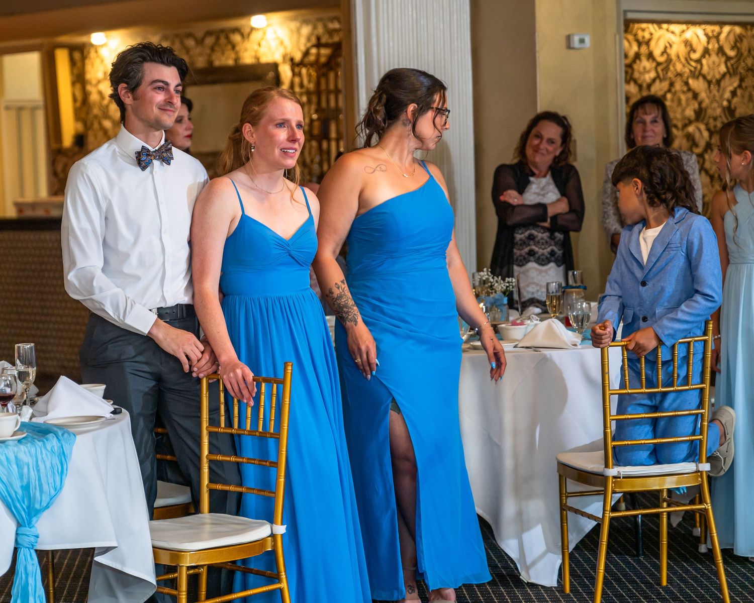 Two bridesmaids in royal blue gowns enter wedding reception hall with groomsman in black tuxedo.