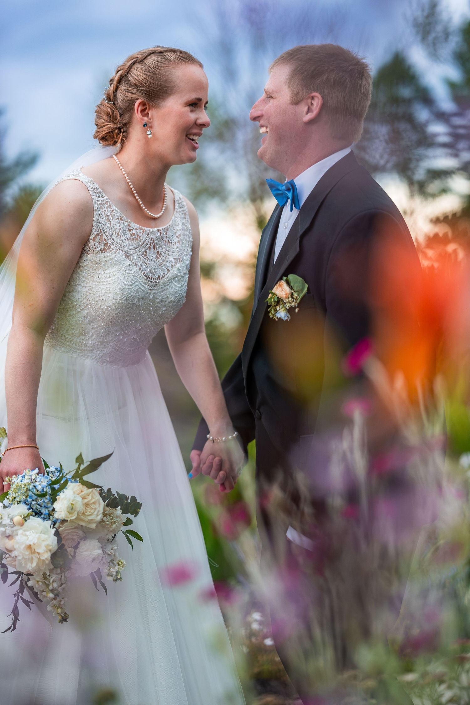 Happy bride and groom hold hands laughing together among colorful flowers at sunset during wedding portraits.