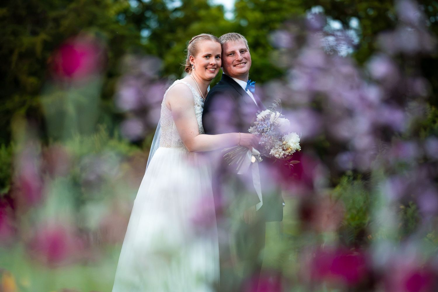 Bride and groom smiling together surrounded by colorful purple and pink garden flowers.