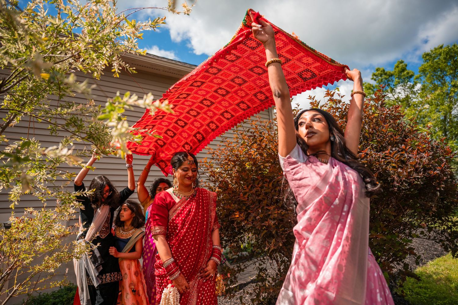 Women in colorful saris hold a red embroidered dupatta overhead during an outdoor South Asian wedding ceremony.