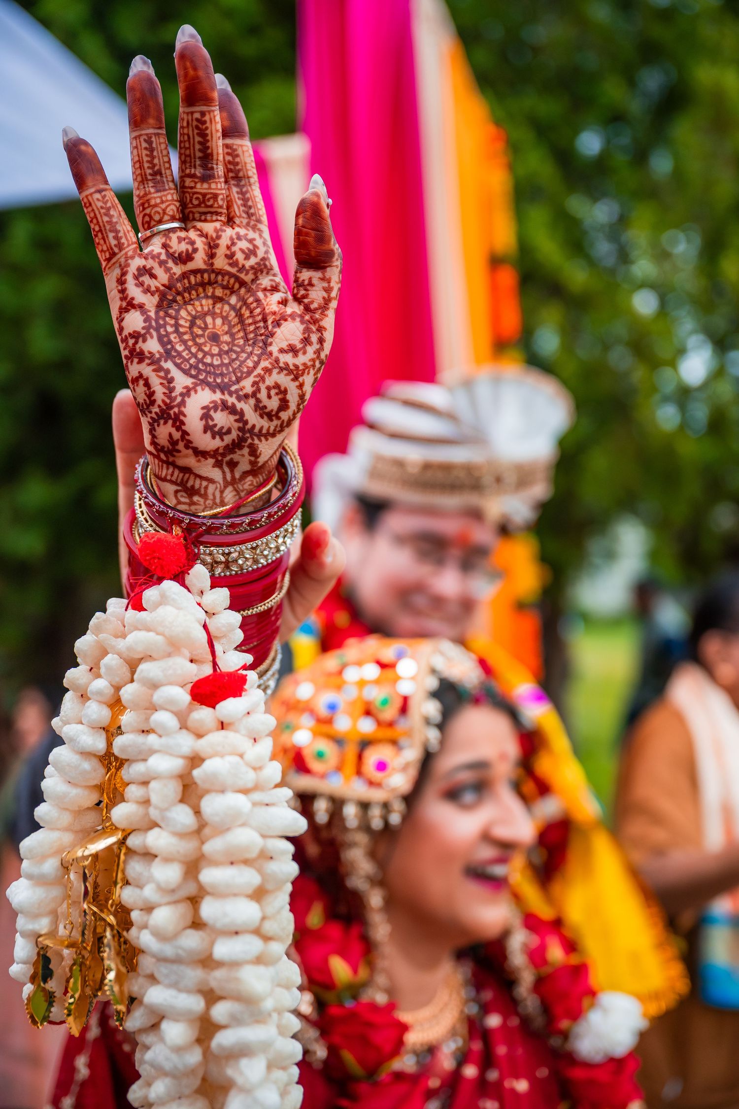 Bride raising henna-decorated hand holding floral garland during Indian wedding ceremony outdoors.