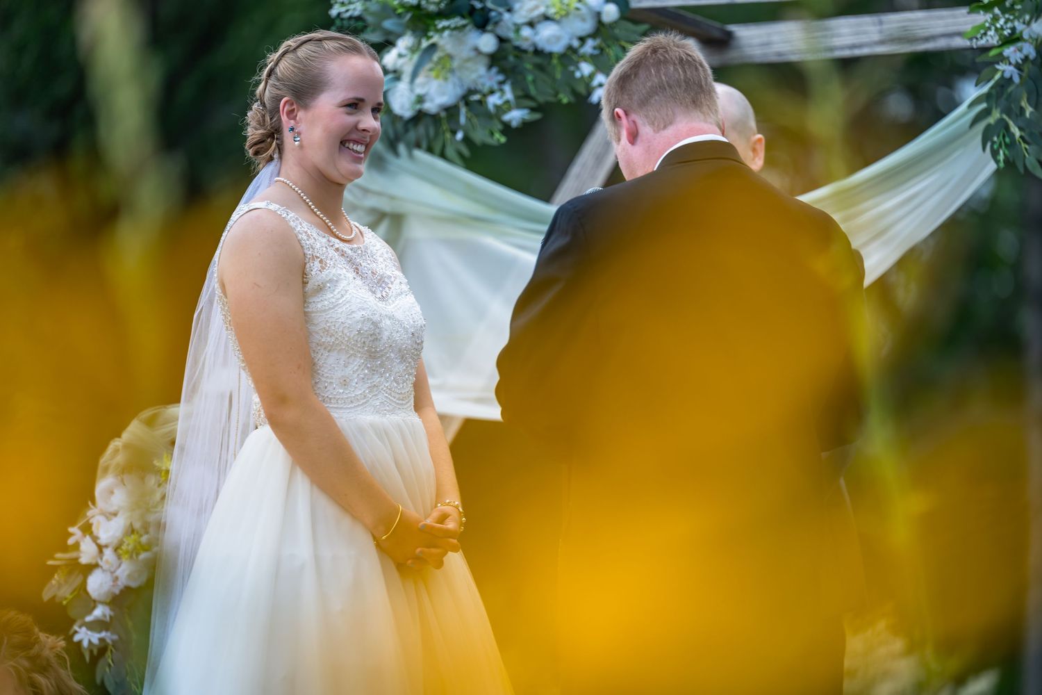 Bride smiling joyfully at groom during outdoor wedding ceremony with floral arch and flowing veil in background.