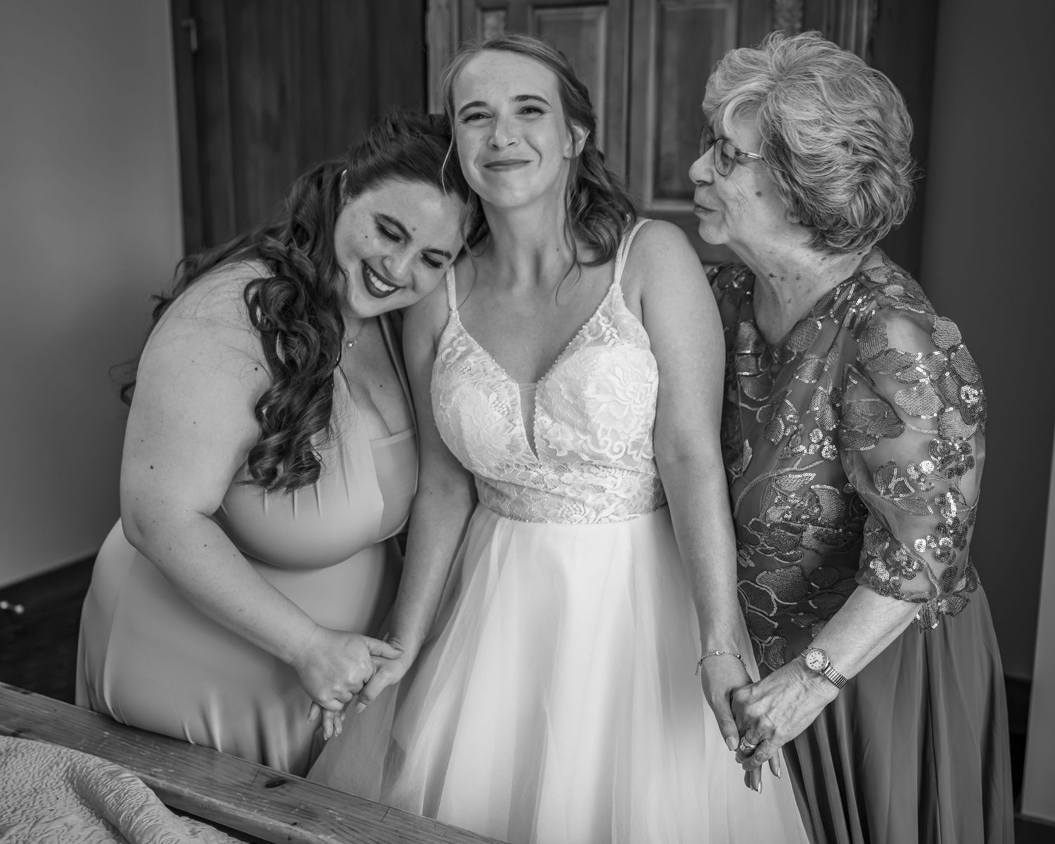 Black and white photo of a bride in a white gown being helped by two women, sharing a joyful moment.