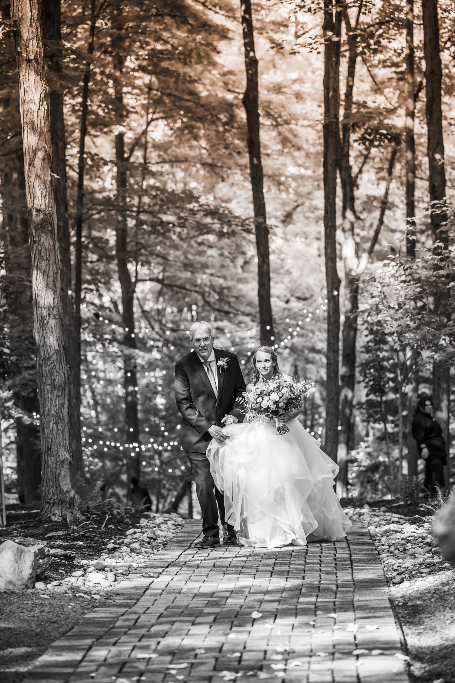 Bride in white gown walks down brick path with escort through sunlit forest wedding venue.