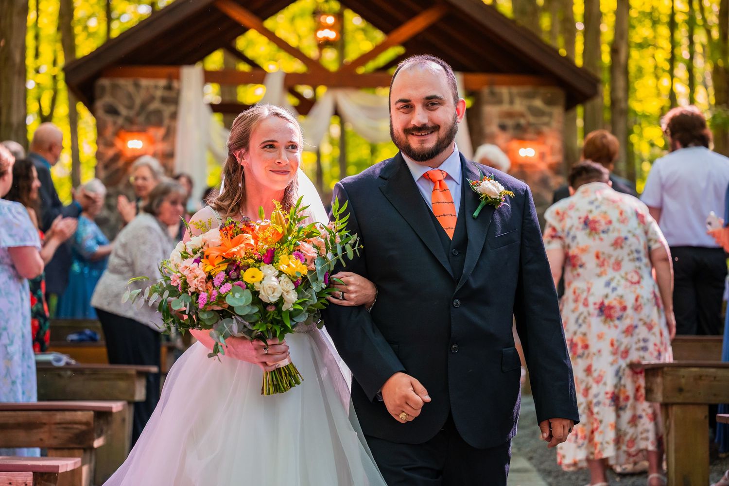 Newlywed couple walks down outdoor chapel aisle surrounded by autumn foliage, bride holding colorful bouquet.