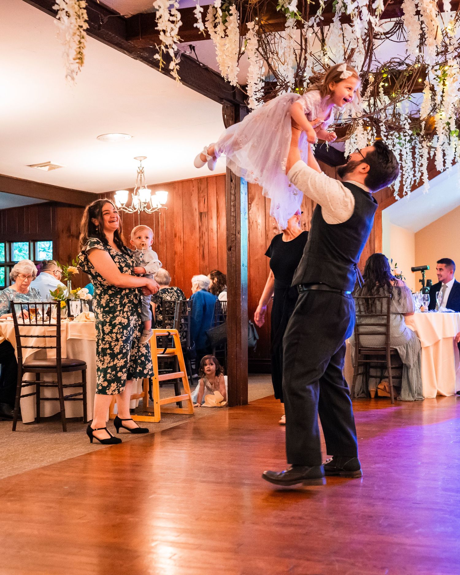 Father joyfully lifts young daughter in pink dress on wedding reception dance floor while mother watches.