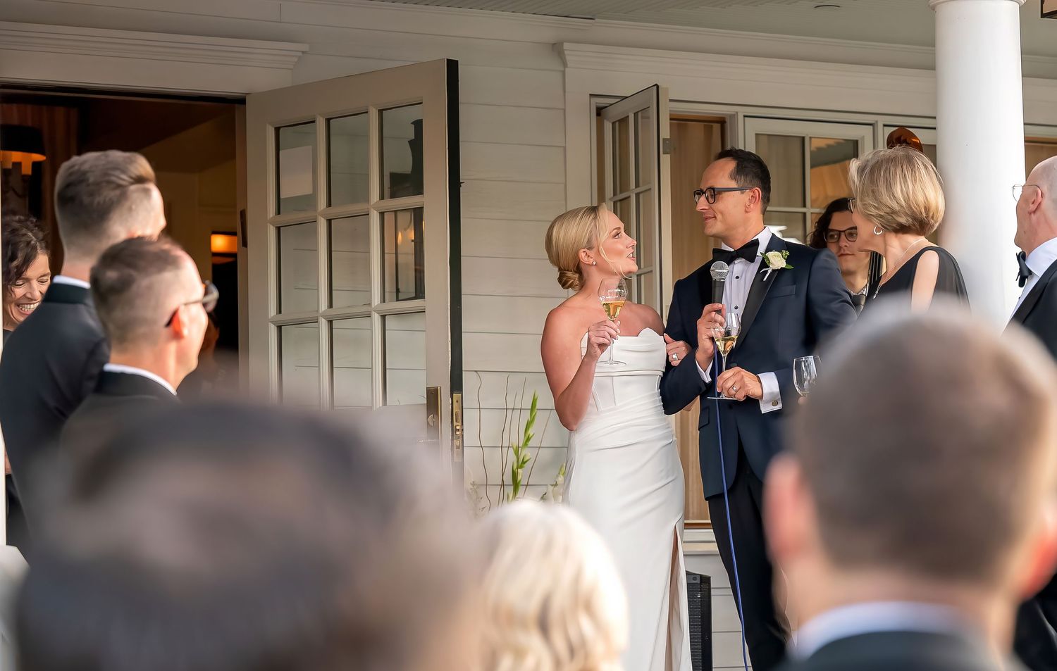 "Glamorous LA couple toasting at their destination wedding in Skaneateles, NY, surrounded by affluent guests on The Krebs restaurant porch"
