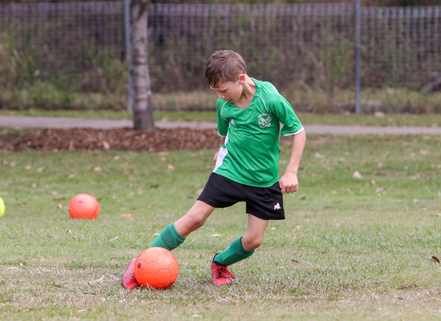 Youth soccer player in green jersey dribbling orange ball on grass field.