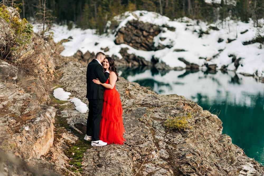 A couple embraces on a rocky cliff overlooking a turquoise alpine lake surrounded by snowy terrain.