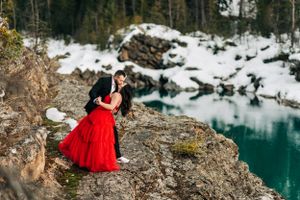 A romantic moment captured on a cliff edge with a stunning turquoise lake and snow-covered landscape below.