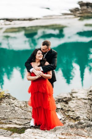 A couple shares an intimate moment by a turquoise alpine lake with snow-covered shores.