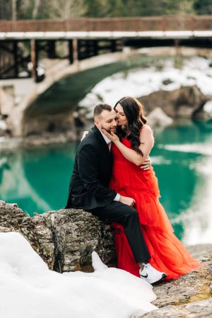 A romantic moment shared on a snowy rock overlooking a turquoise lake with a bridge in the background.