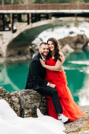 A couple poses on a snowy rock with a scenic bridge and turquoise waters in the background.
