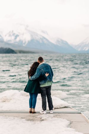 A couple stands close together on a dock overlooking a choppy mountain lake on a cloudy day.