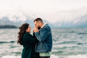 A romantic winter engagement photo by a lake with snow-capped mountains in the background.