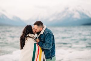 Couple embracing by mountain lake wrapped in colorful striped blanket during winter.