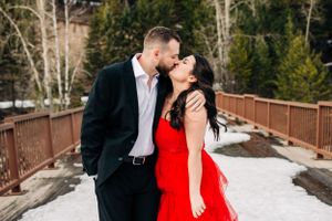 Romantic winter engagement photo on wooden bridge with snow and evergreen trees.
