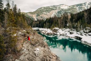 Aerial view of couple in red standing on rocky overlook above turquoise river in snowy mountains.