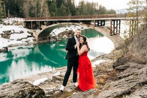 A couple poses by a stone bridge over turquoise water in a snowy mountain landscape.