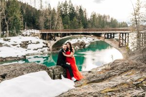 Two people embrace near a concrete bridge spanning bright turquoise waters and snowy banks.