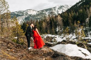 A couple stands on snowy terrain with mountains and evergreen forest in the background.