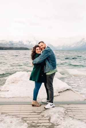 A couple embraces on a snowy dock with mountains and choppy waters behind them.