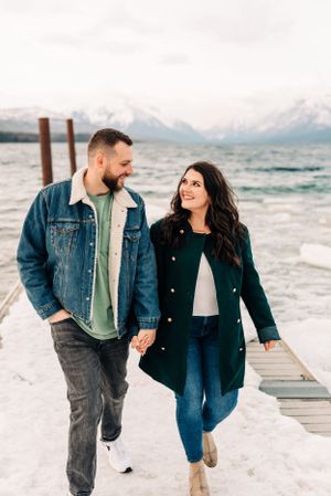 A couple walks hand in hand on a snow-covered dock with mountain views.