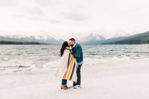 A couple shares an intimate moment on a snowy beach with mountains in the background.