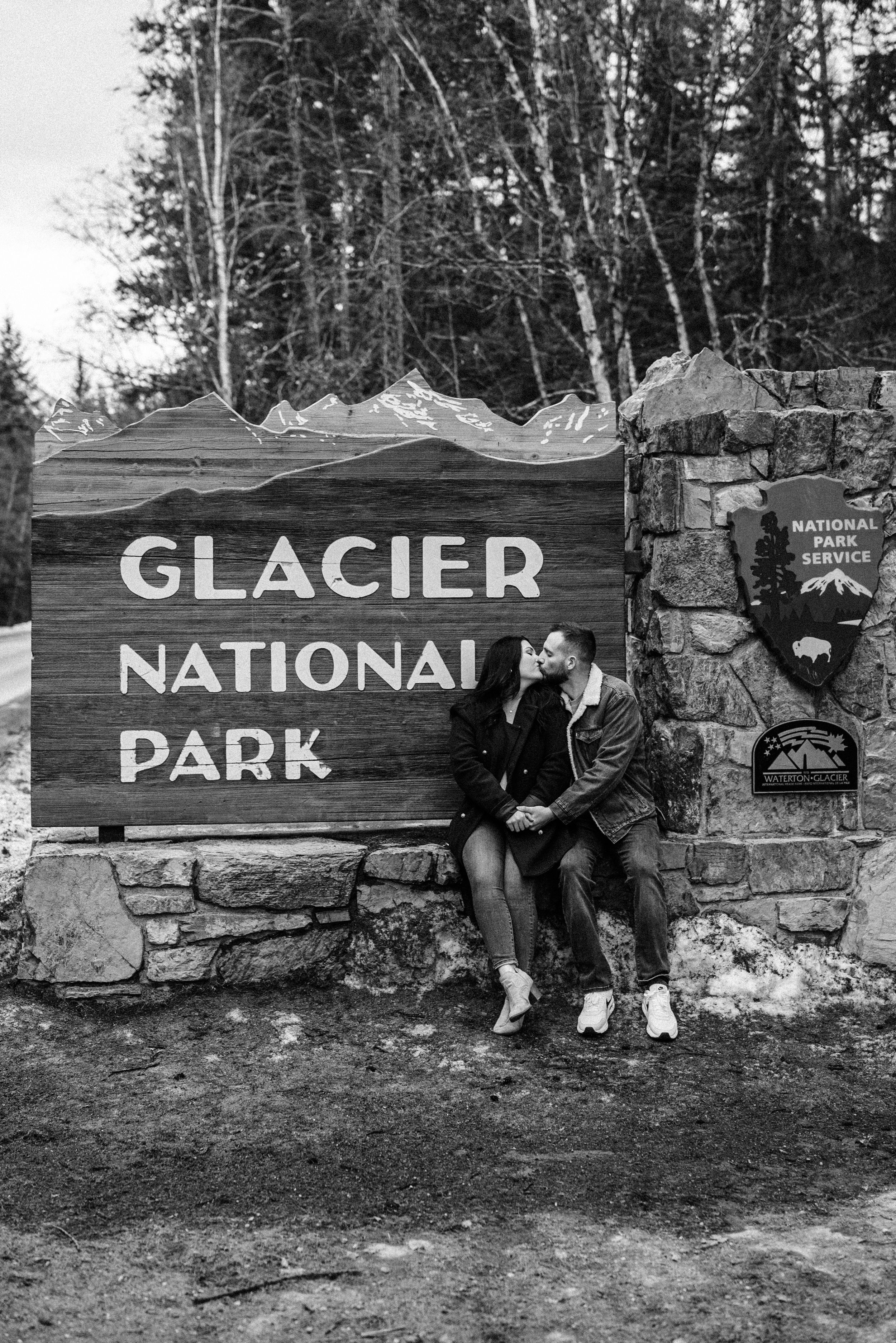 Black and white photo of visitors sitting on stone wall beneath Glacier National Park entrance sign.