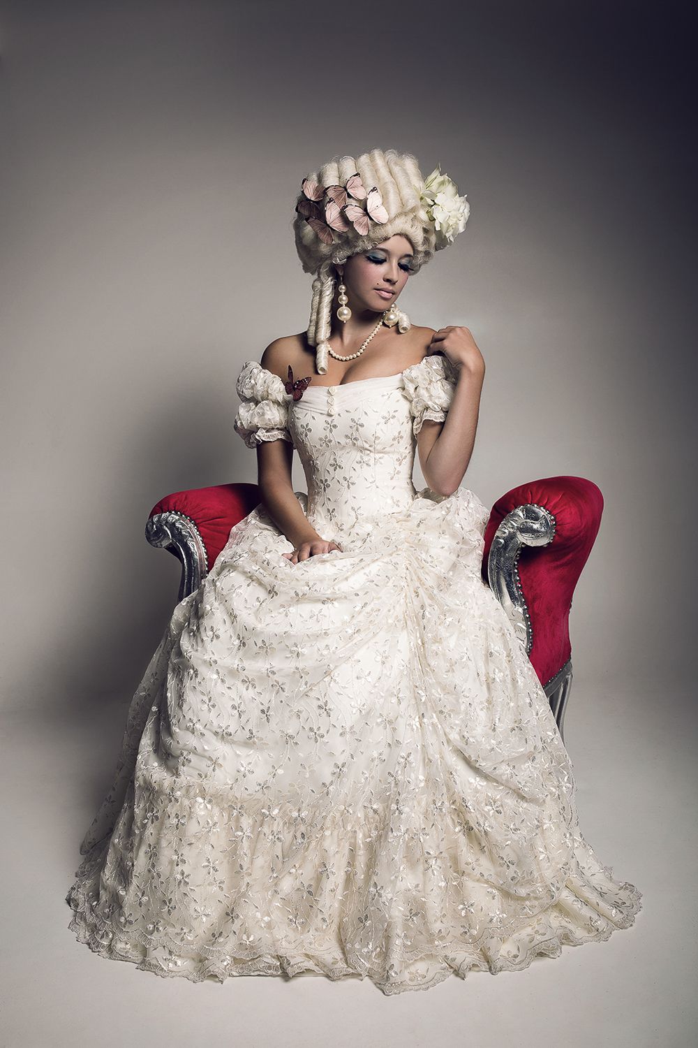 Woman in baroque white ball gown and powdered wig seated on red velvet chair.