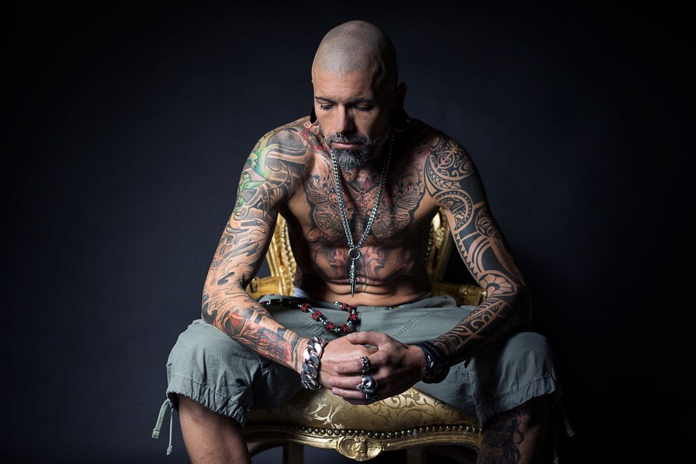 Heavily tattooed bald man sitting shirtless on golden chair in dark studio.