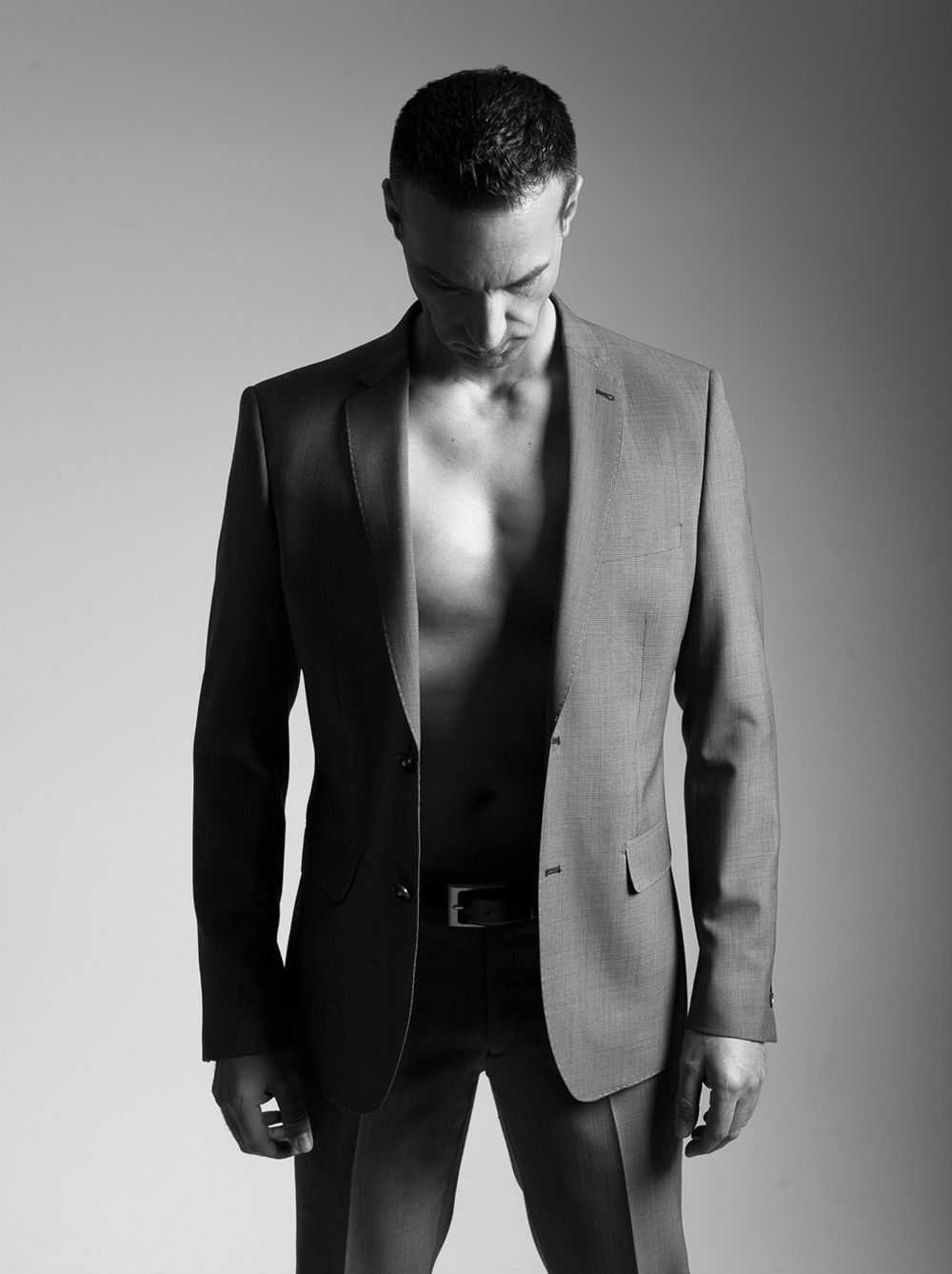 Athletic man in black blazer with open chest posing in dramatic black and white studio portrait.