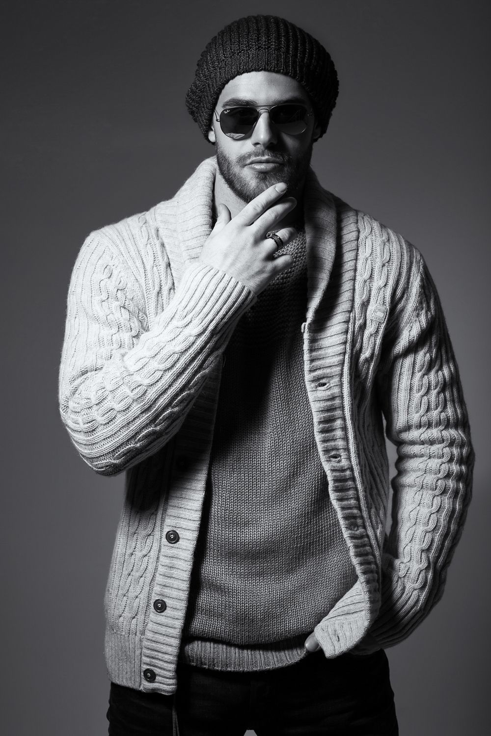 Stylish bearded man in knit cardigan and beanie hat wearing sunglasses in black and white portrait.