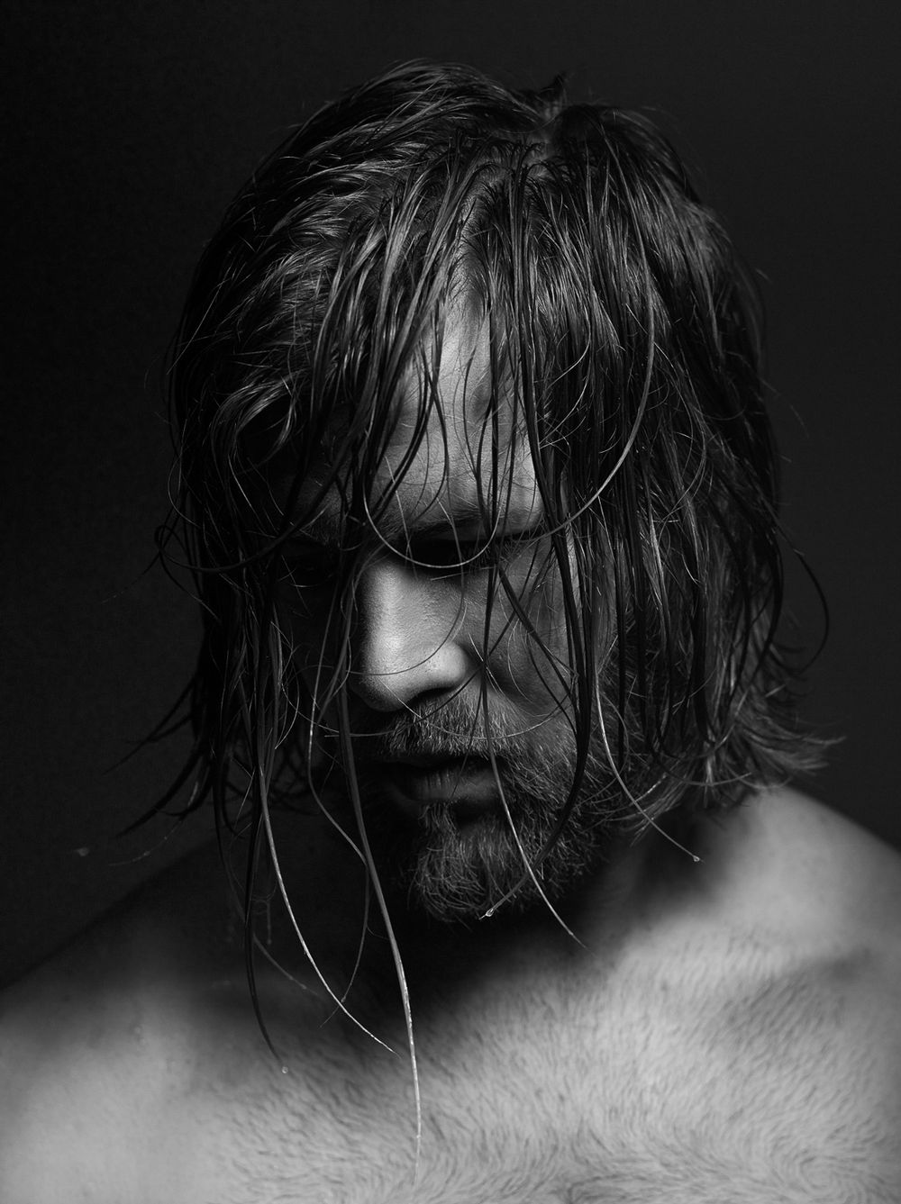 Dramatic black and white portrait of a long-haired man with wet hair and beard looking downward.
