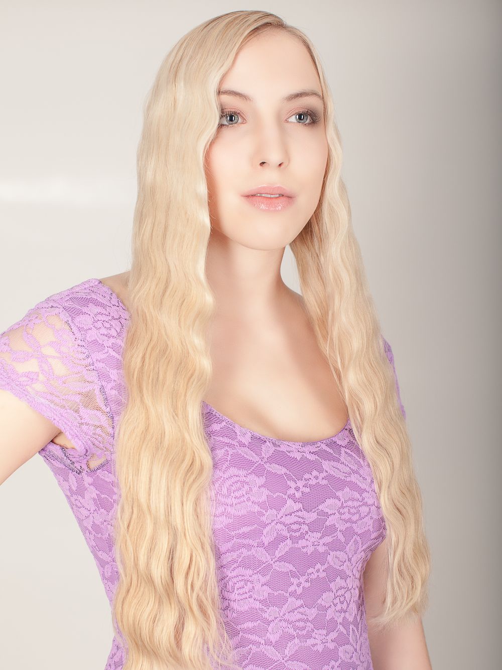 Young blonde woman with long wavy hair wearing a purple lace top in a studio portrait.