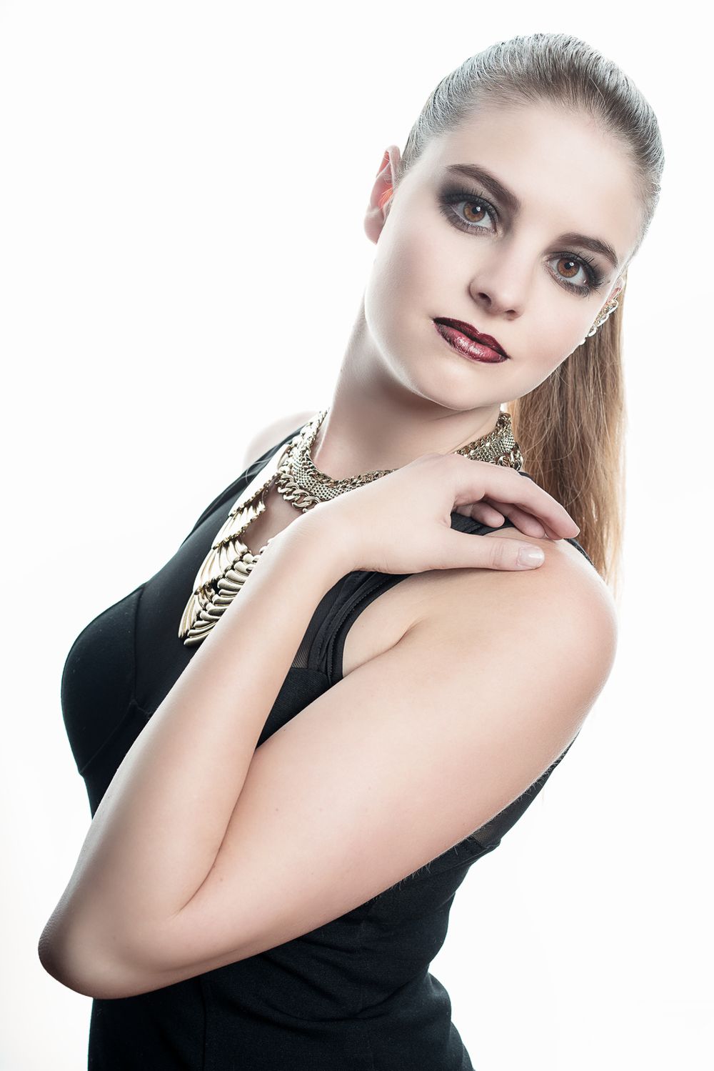 Elegant young woman in black dress with gold statement necklace and bold makeup on white background.